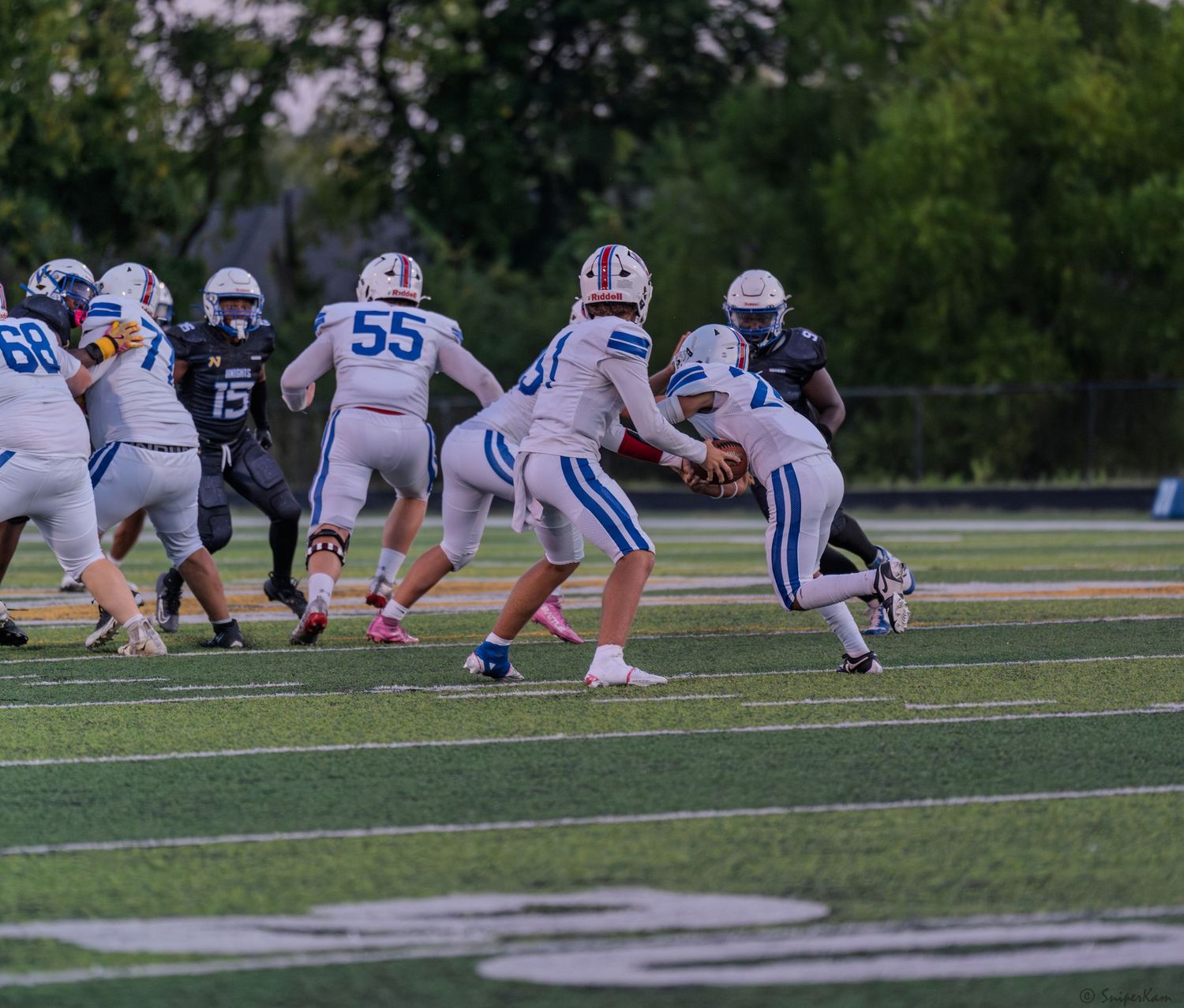 Football players in blue uniforms execute a handoff play during a daytime game.
