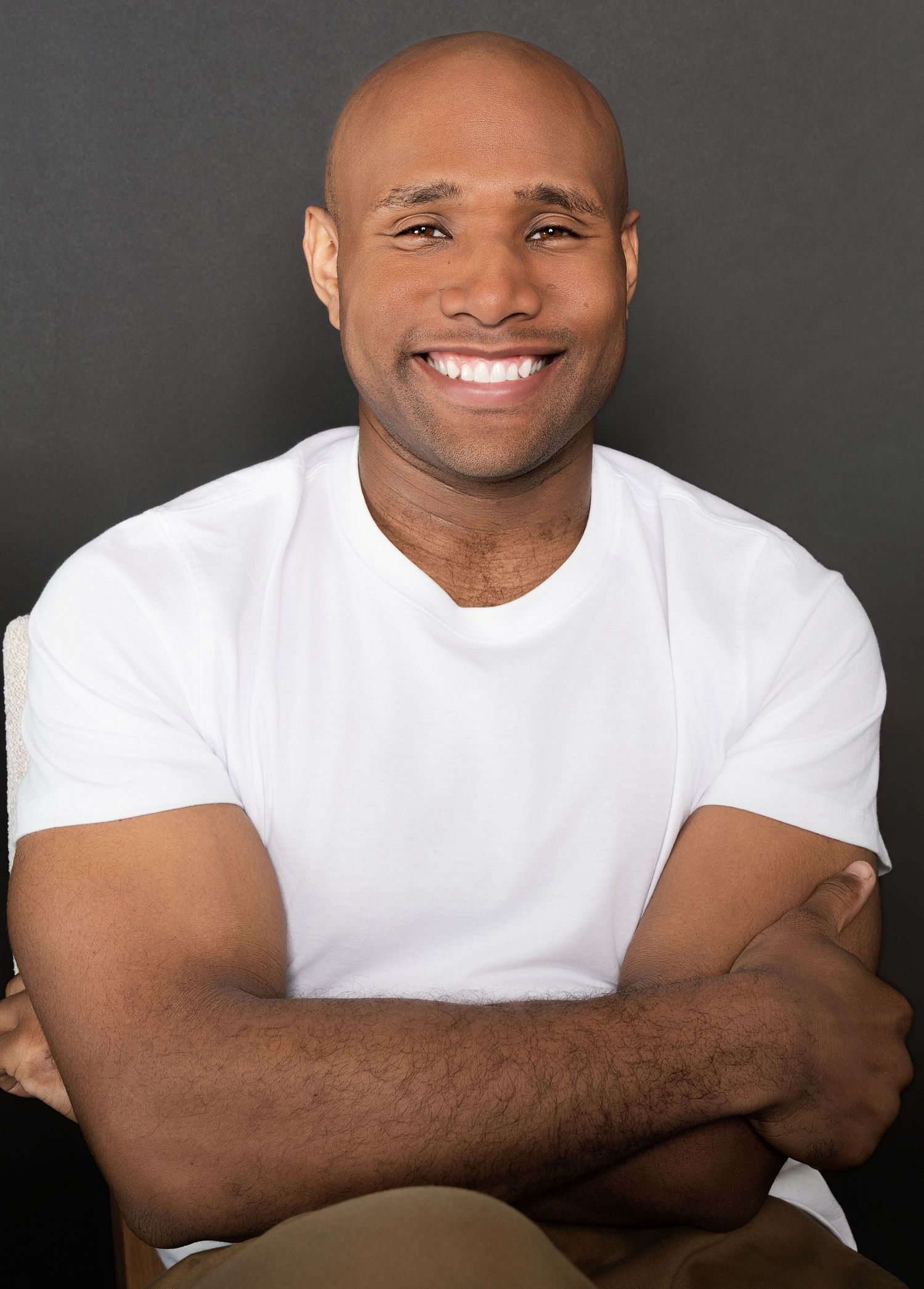 Happy bald Black man in white t-shirt smiling with arms crossed in color portrait against dark gray background.