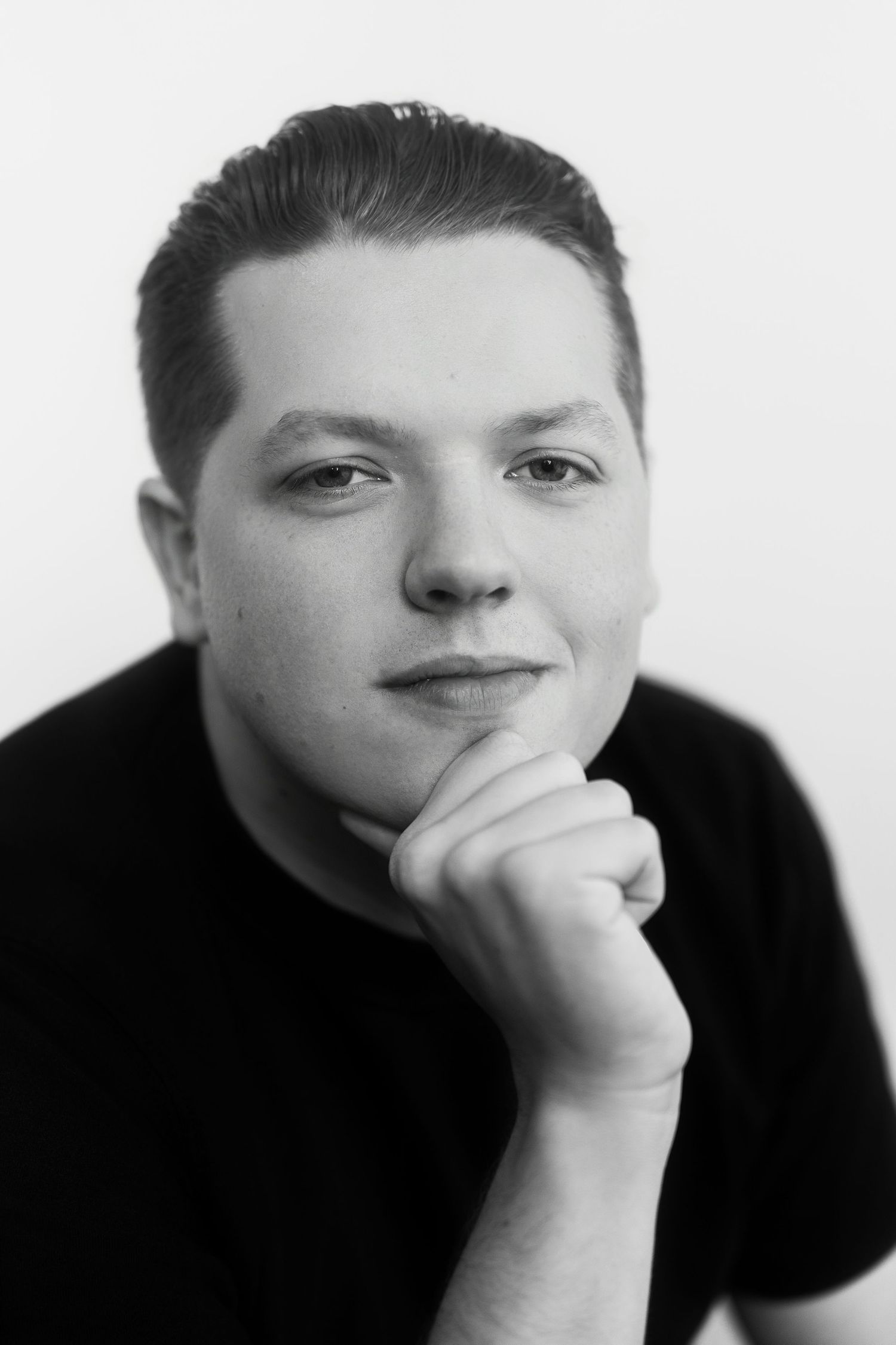 Black and white portrait of young man in black t-shirt resting chin on hand against white background.