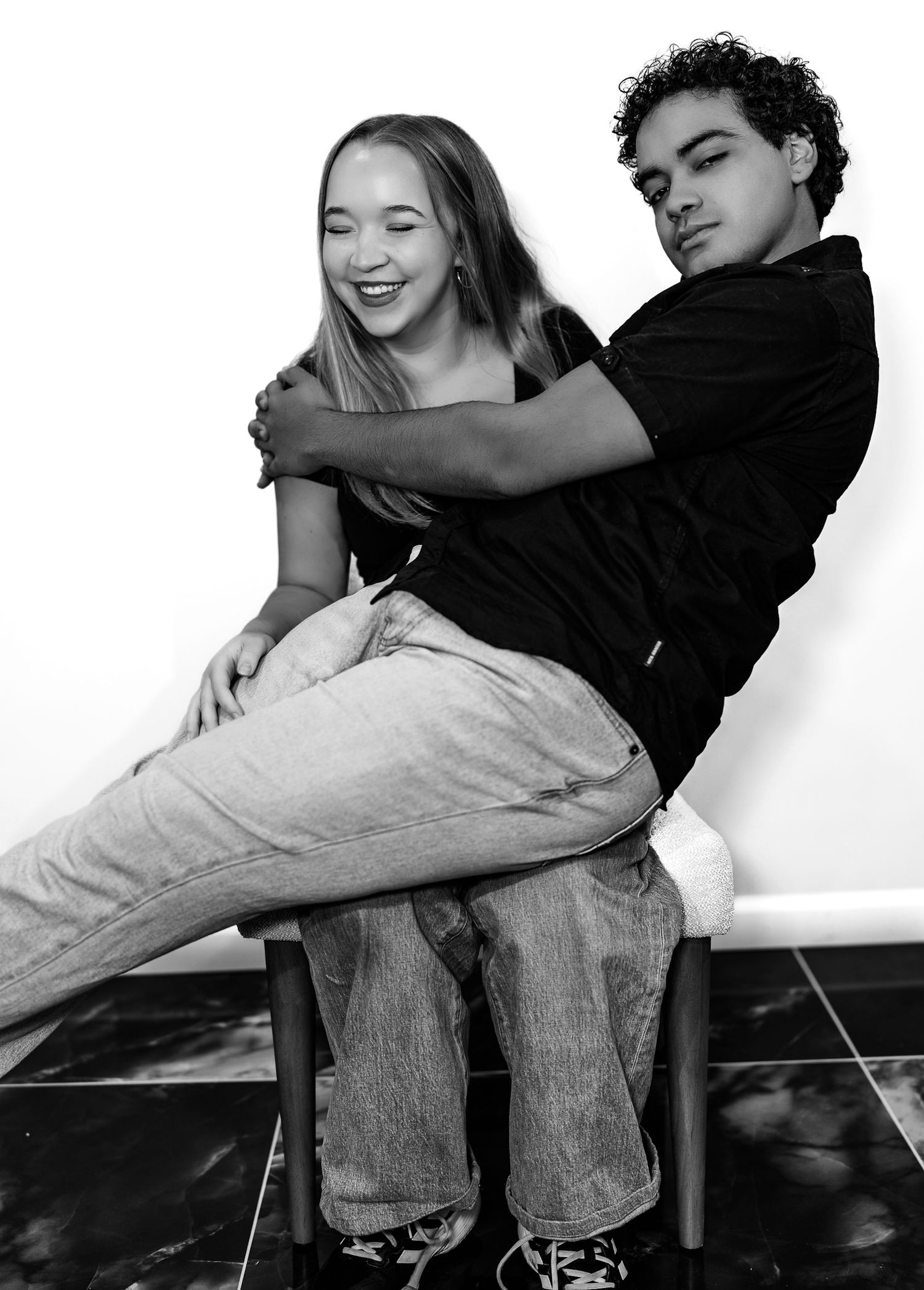 A smiling young woman sits on a bench while a young man leans against her in black and white.