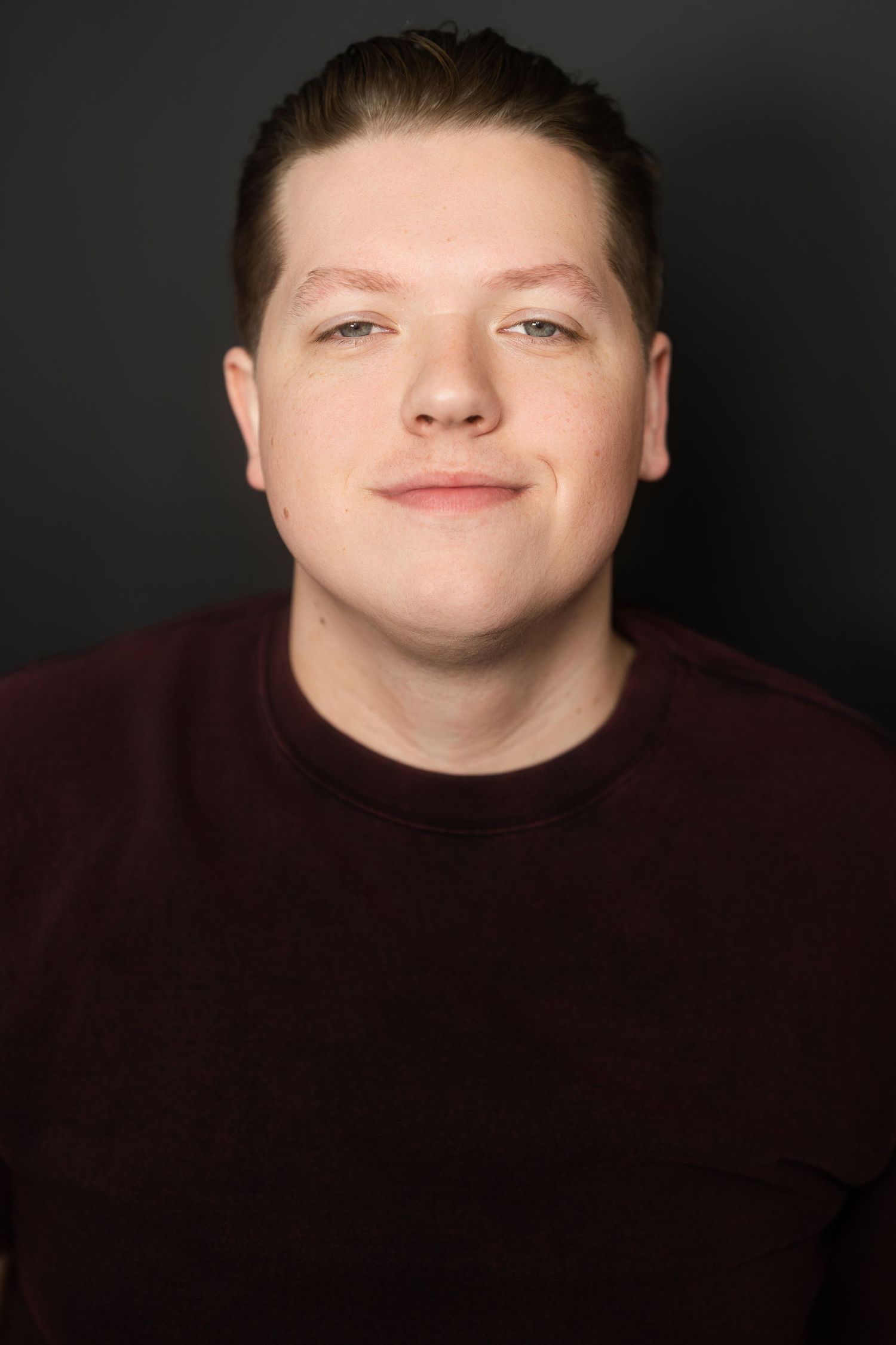 Young man with short hair wearing a dark maroon sweater poses for a professional headshot against a gray background.