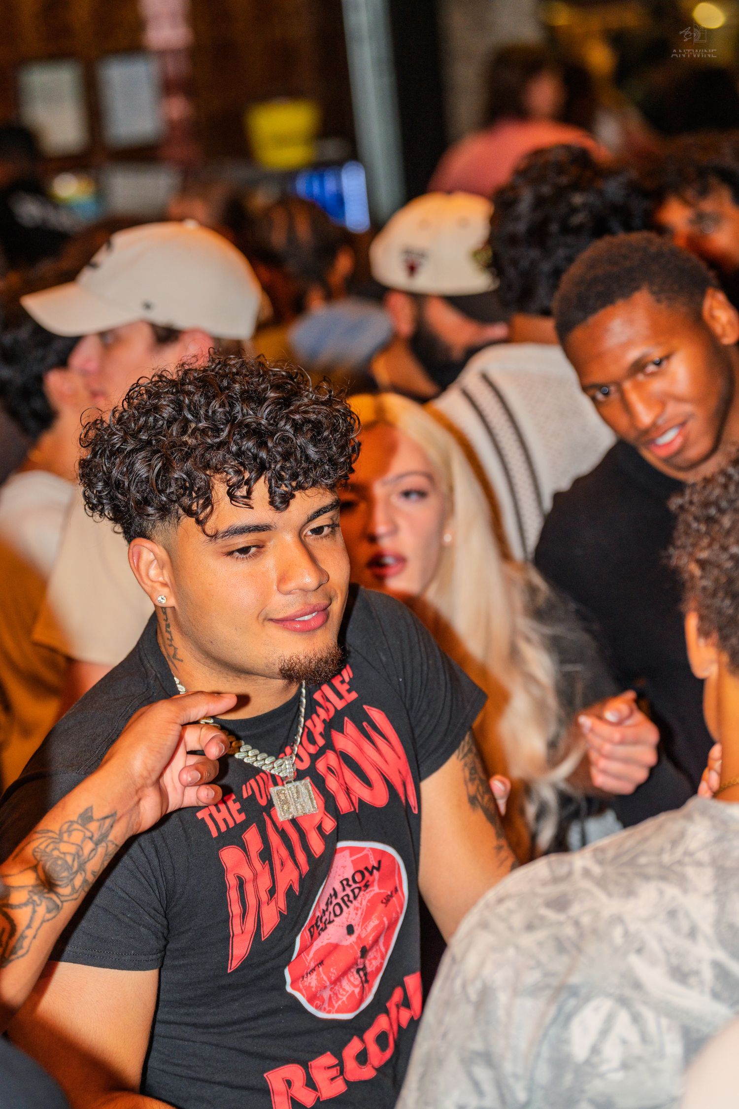 Young man with curly hair and tattoos wearing a Death Row Records shirt at a crowded nightlife event.