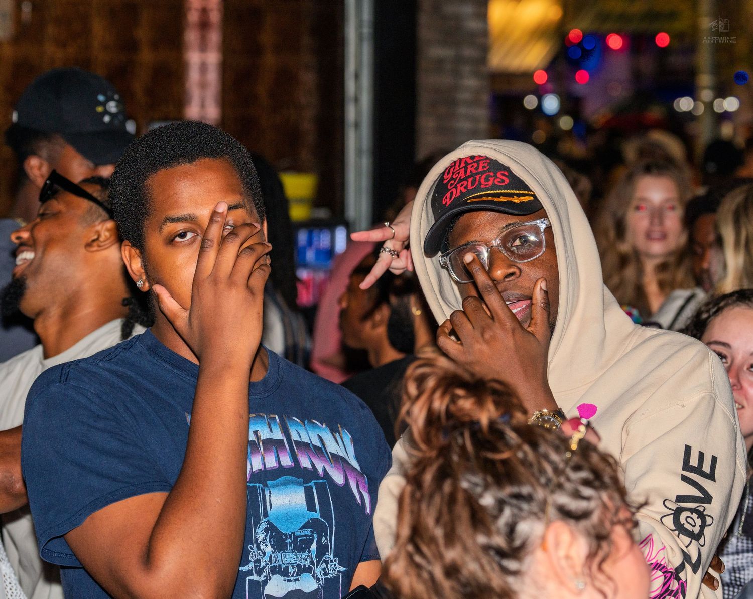 Two young men covering their faces at a lively outdoor nighttime event.