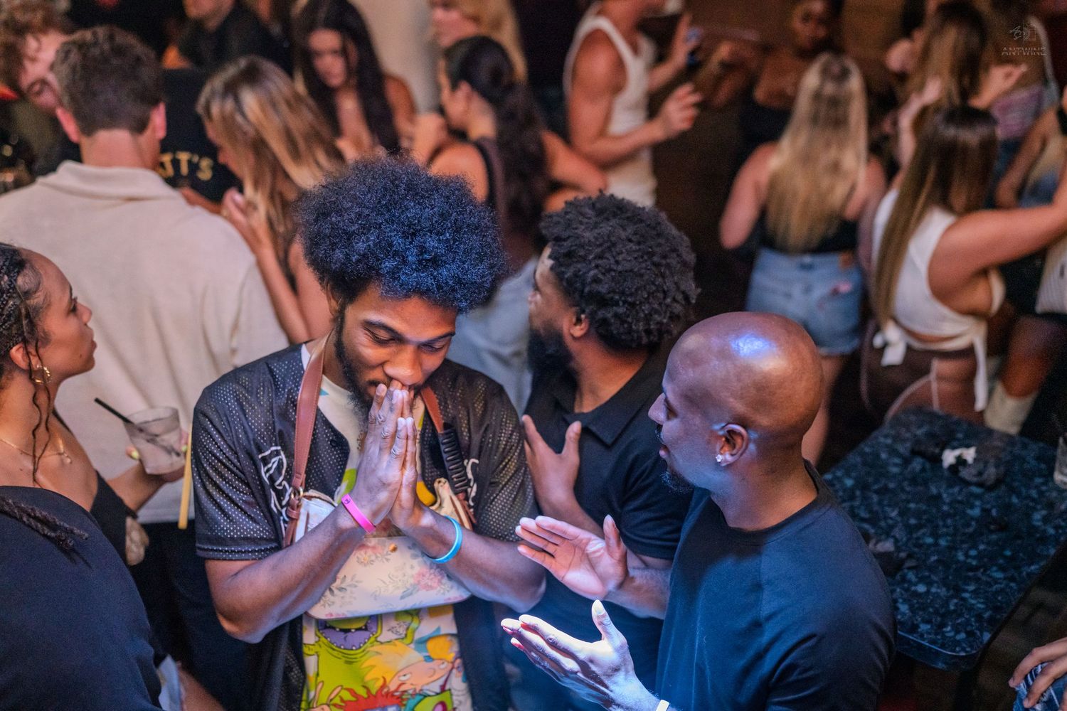 Young man with afro hair praying hands gesture at crowded nightclub party event.