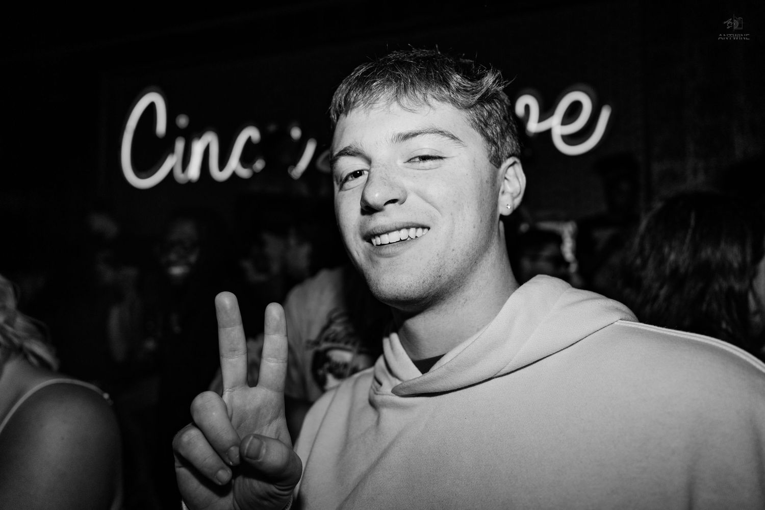 Young man smiling and making peace sign at a nightclub with neon signs in background.