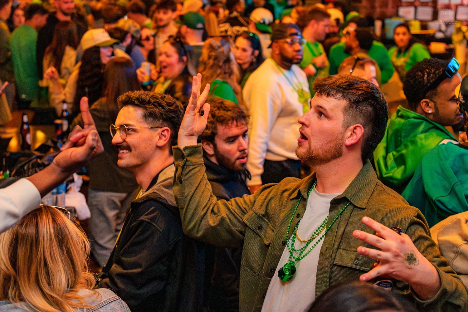 Men in green outfits raising hands and gesturing at a packed St. Patrick's Day celebration event.