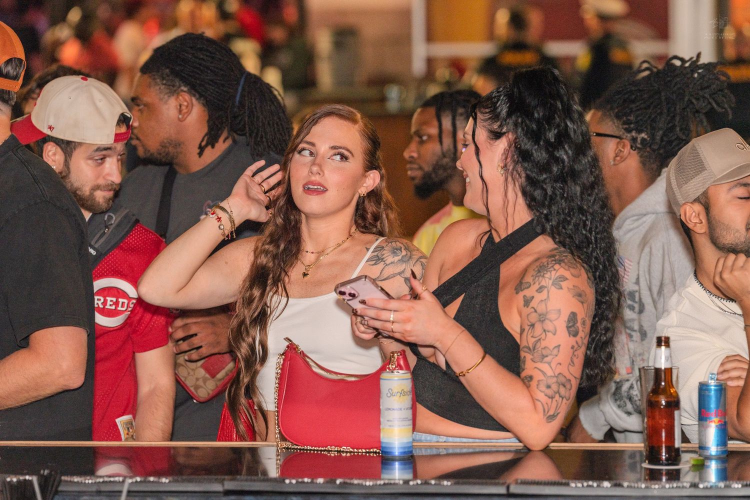 Young woman with red bag chatting with tattooed friend at a crowded bar with beer bottles nearby.