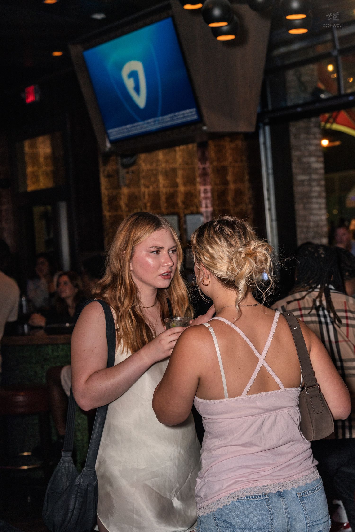 Two women conversing at a bar, one in white dress holding a drink, other in pink strappy top.