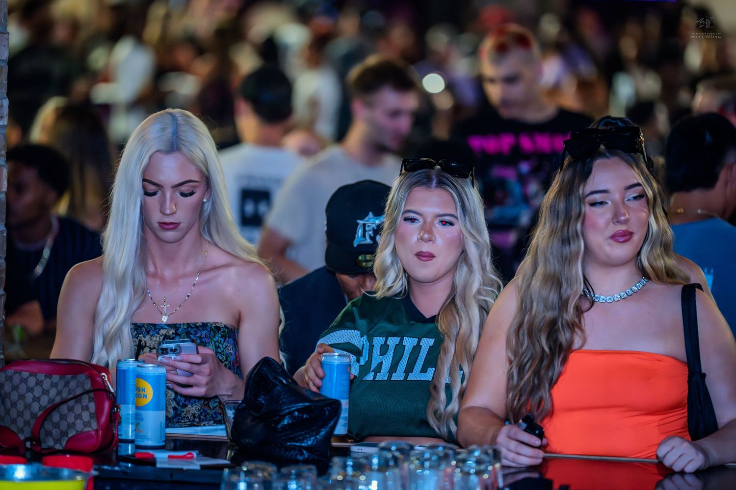 Three women sitting at a bar in a nightclub, looking at phones amid a large crowd.