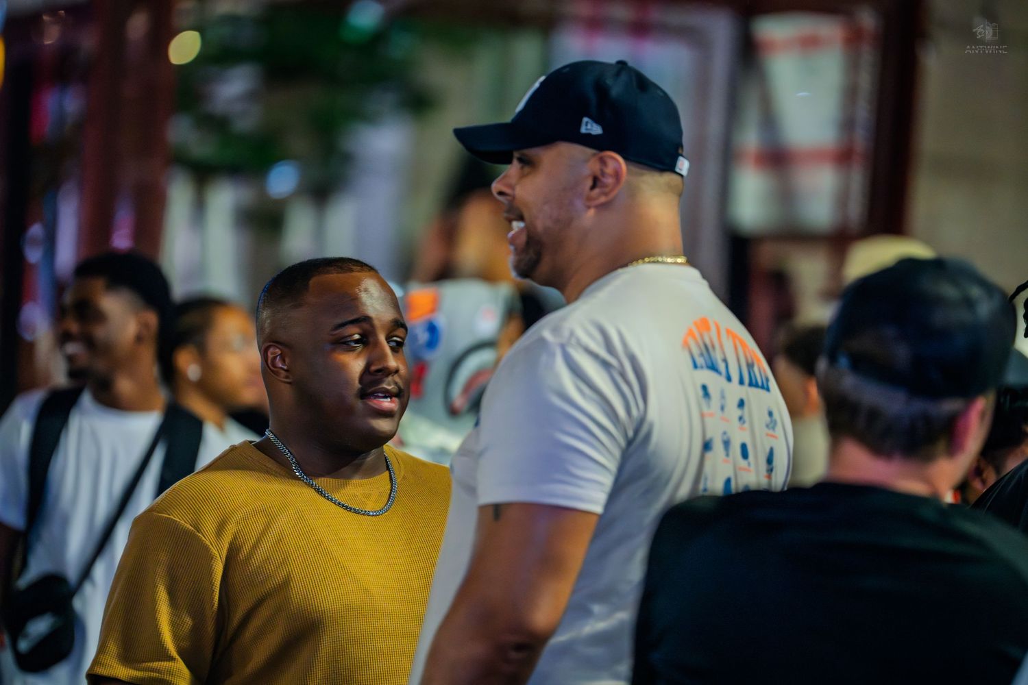 Two men conversing at an outdoor nighttime event, one in yellow shirt.