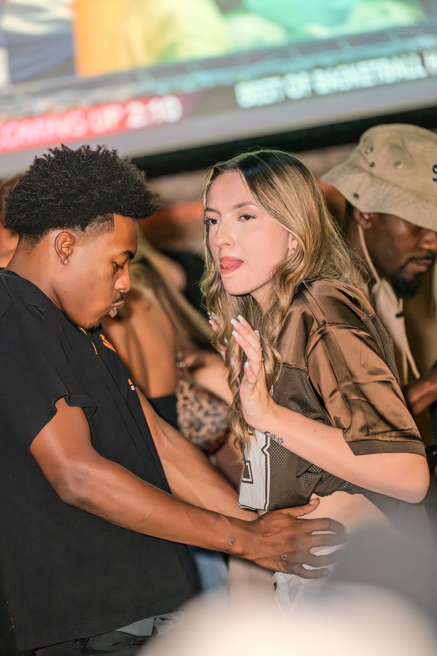 Young woman in brown jacket talking with a man at a crowded nightclub event.