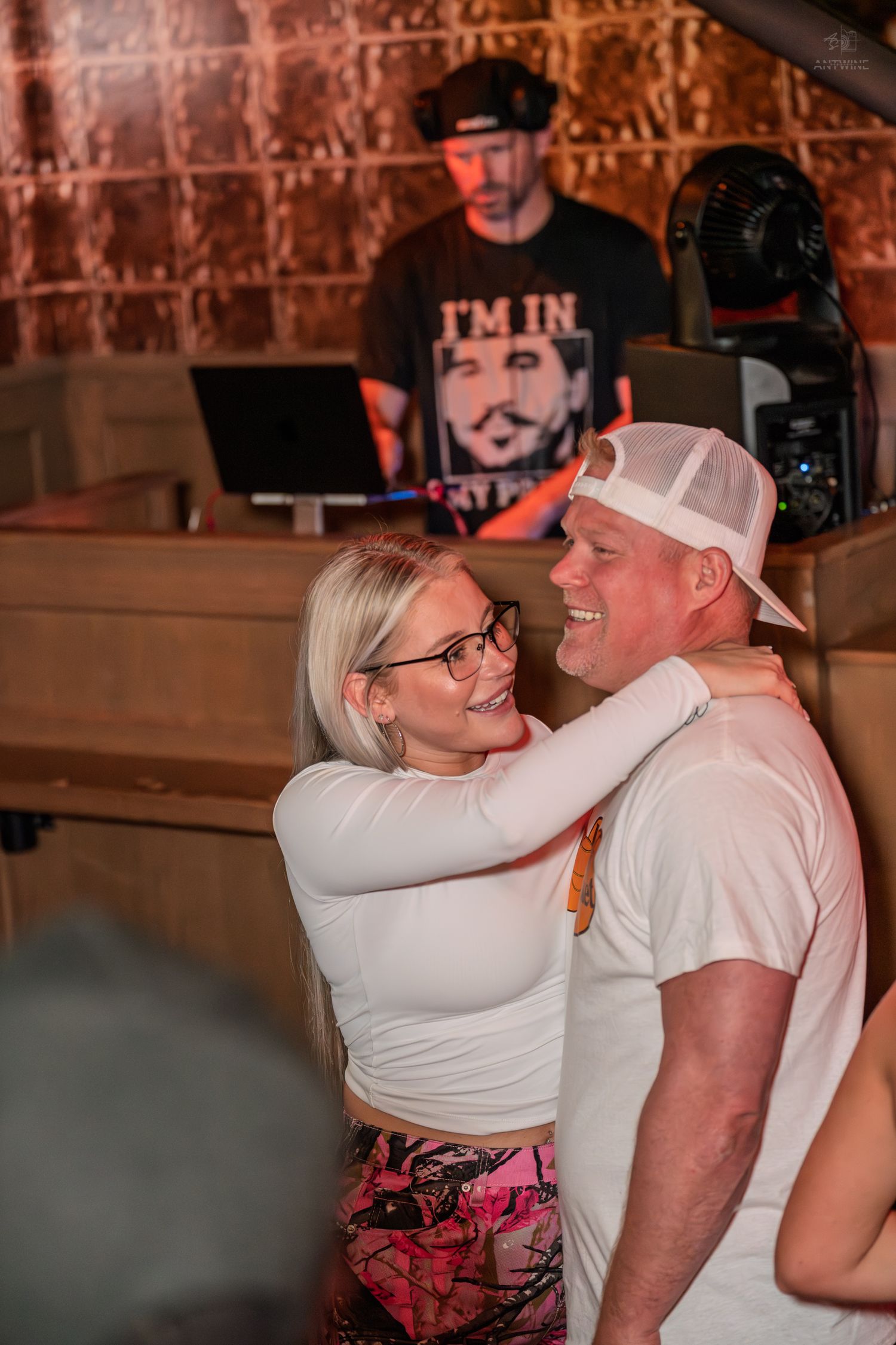 Couple smiling and embracing on a dance floor with a DJ performing behind them.