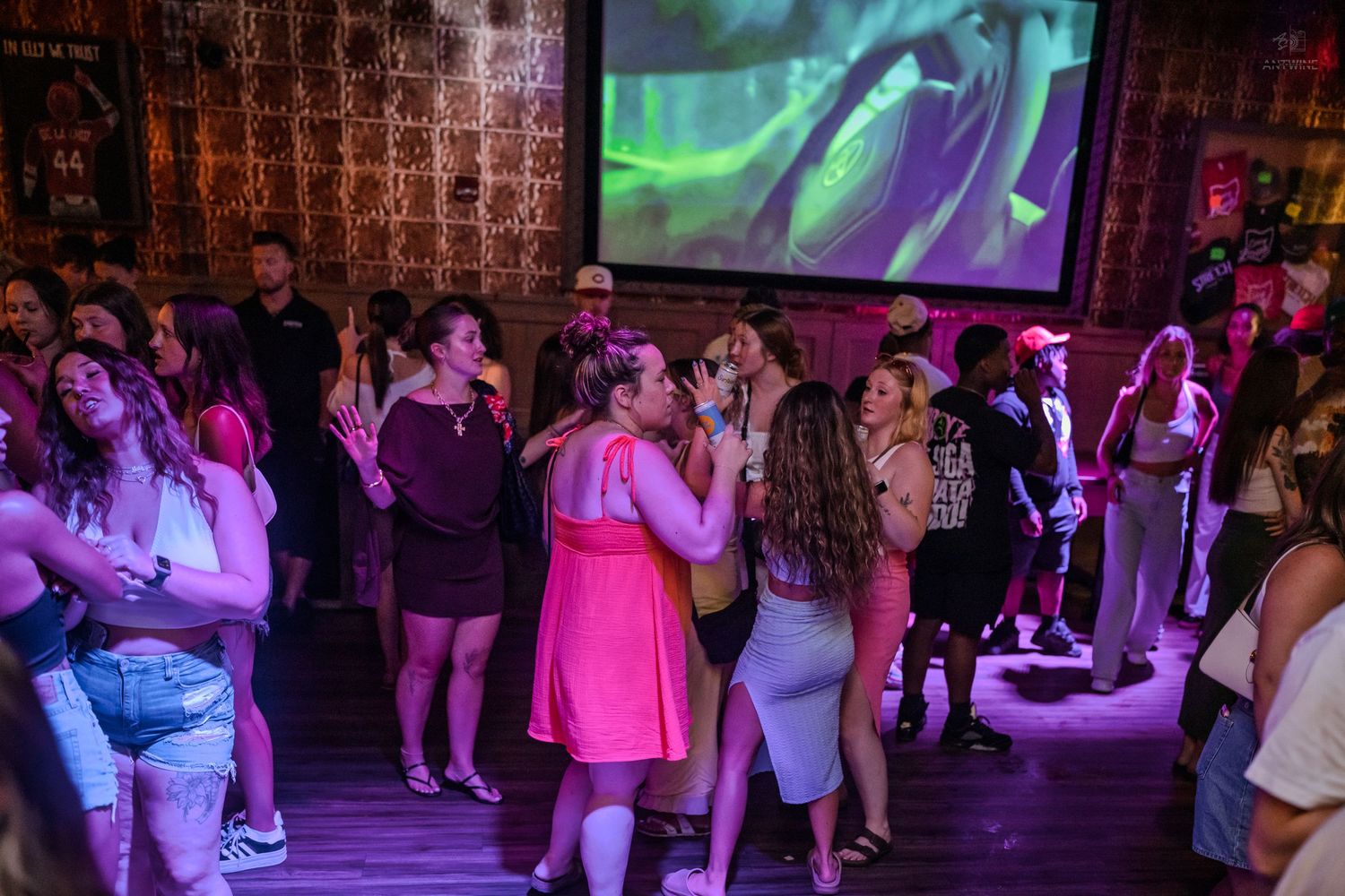 Energetic crowd singing and dancing in a nightclub with a large projection screen behind them.