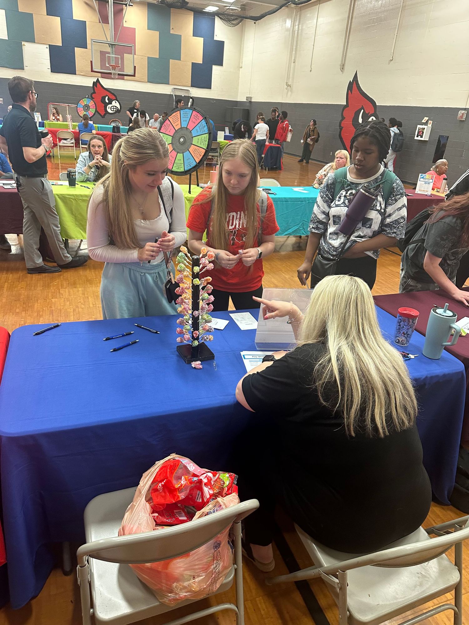 Students and adults interact at a colorful educational fair in a school gymnasium with Cardinal mascot decorations.