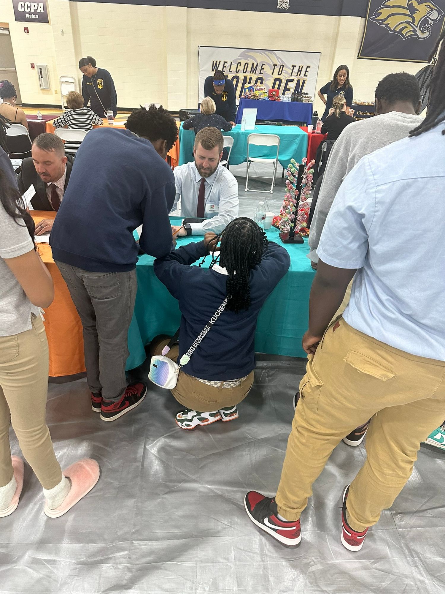 Students gather around a teal booth at a school career fair inside a gymnasium with gold and blue decor.
