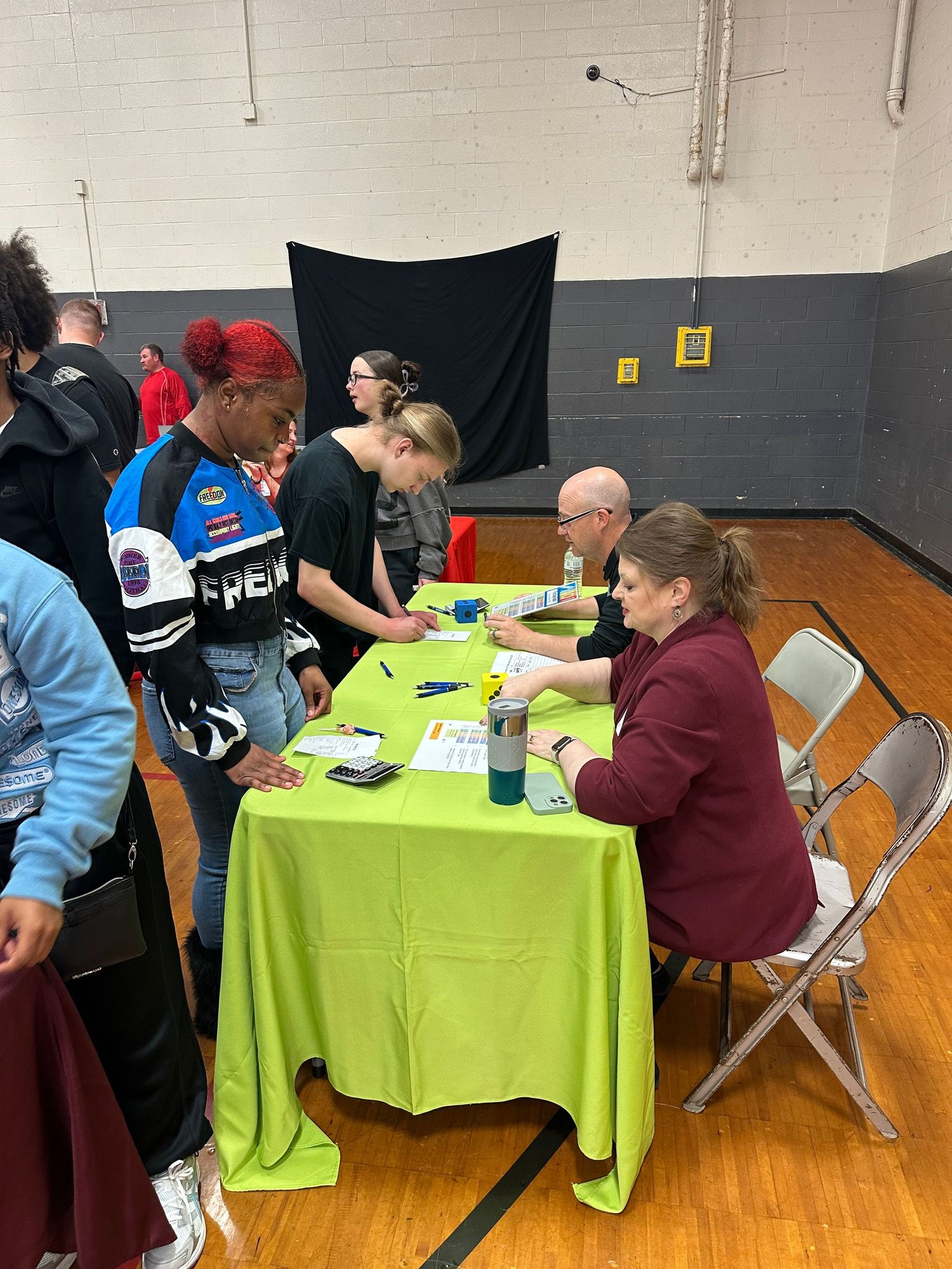 Students stand at a bright green table in a gym, interacting with seated adult representatives at a school event.