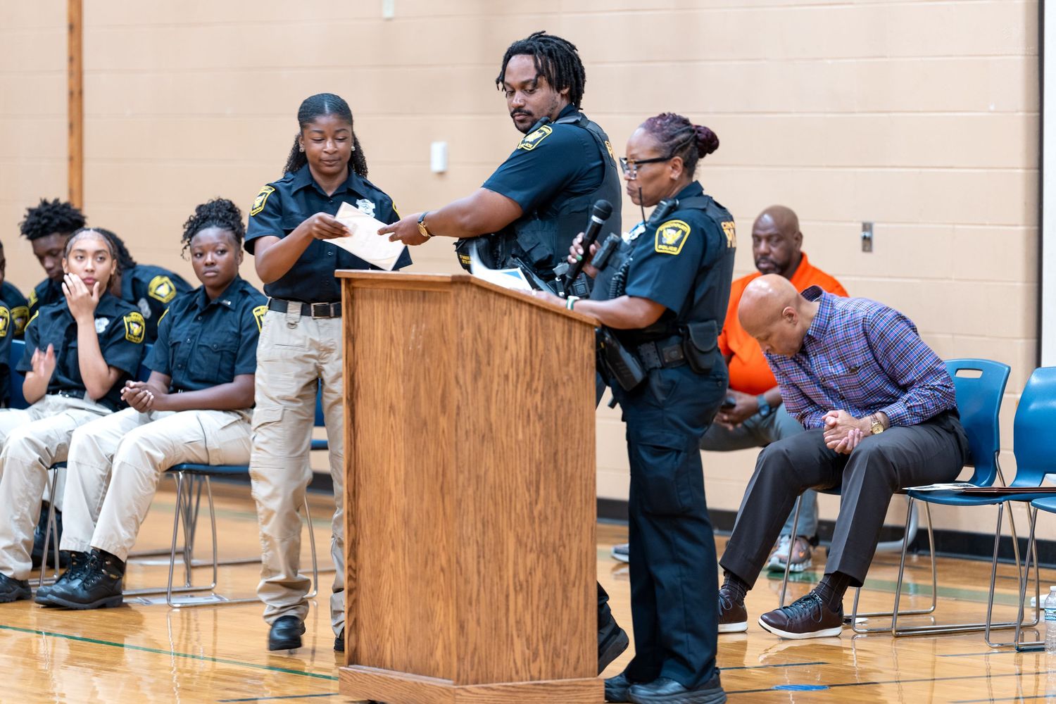 Law enforcement officers address community members at a public meeting held in a gymnasium.
