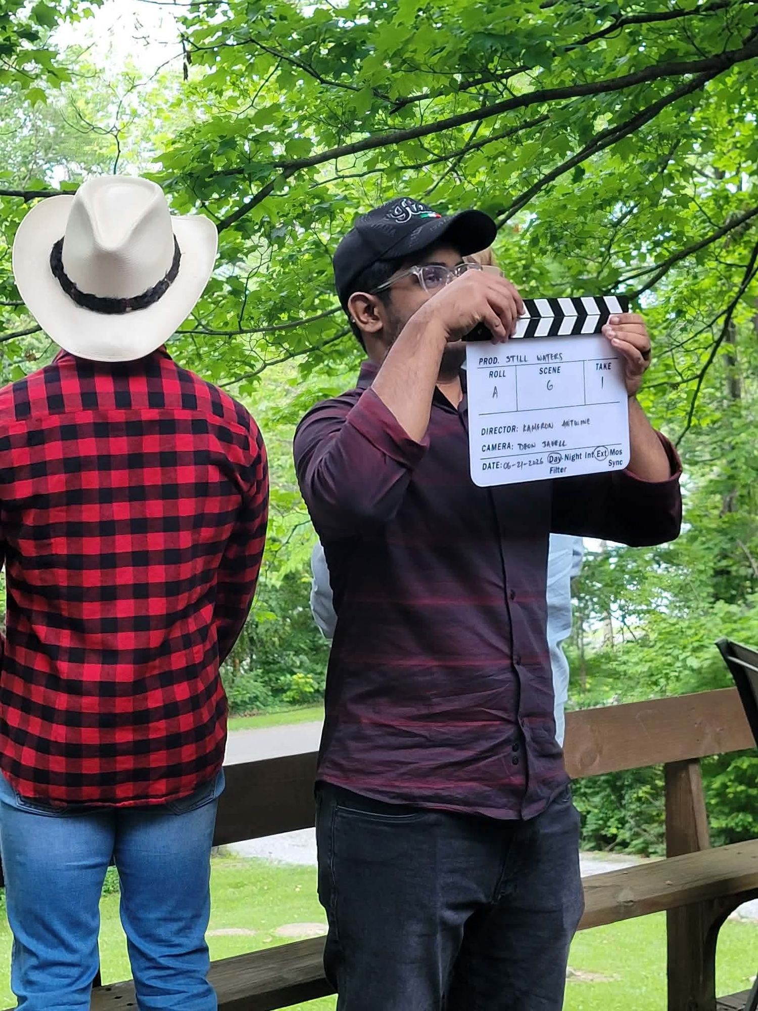Film director holding clapperboard next to actor in red plaid shirt and cowboy hat on outdoor wooden deck.