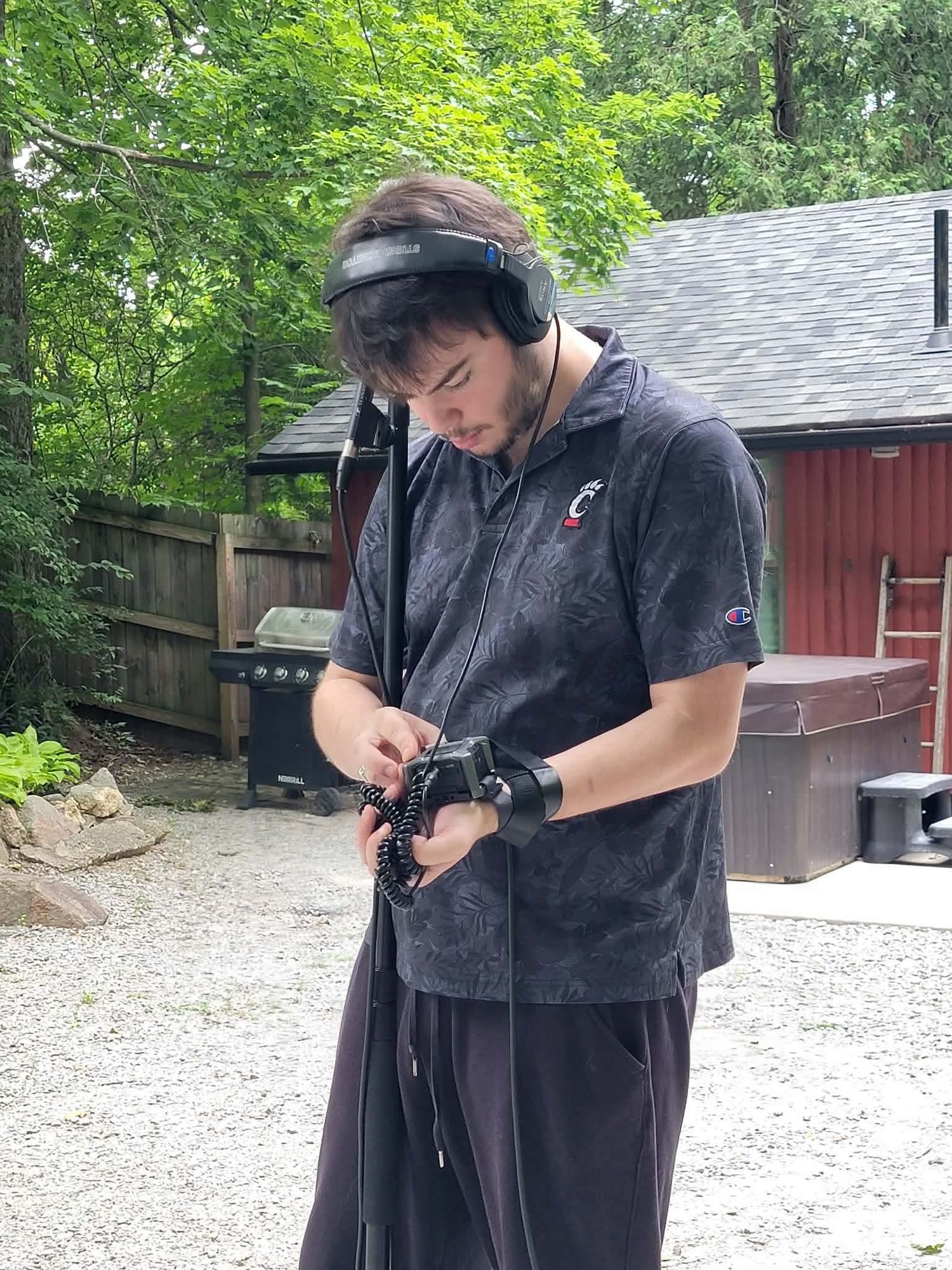 A young man wearing headphones and a Champion hoodie adjusting audio equipment outdoors.