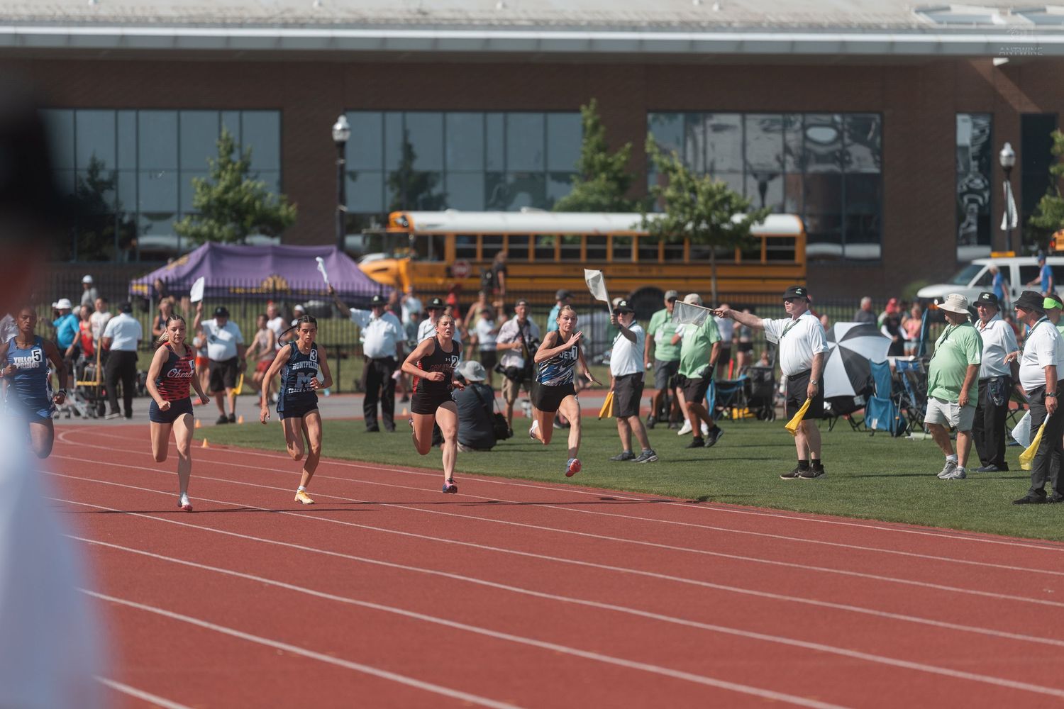 Student runners compete in a distance race on a red outdoor track at a high school track and field meet.