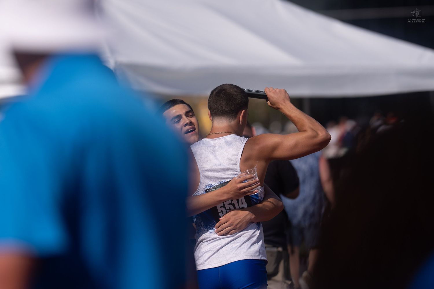 Two athletes share a celebratory embrace at an outdoor track and field competition on a bright sunny day.