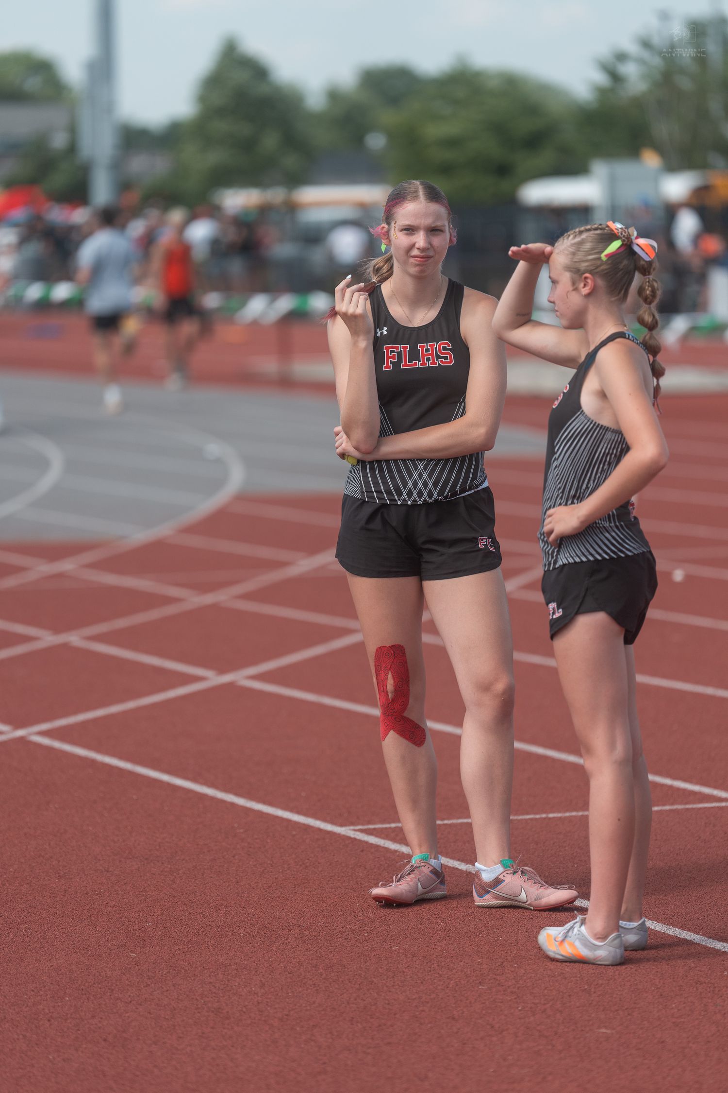 Two young female track athletes in black uniforms stand talking on a red track at a meet.