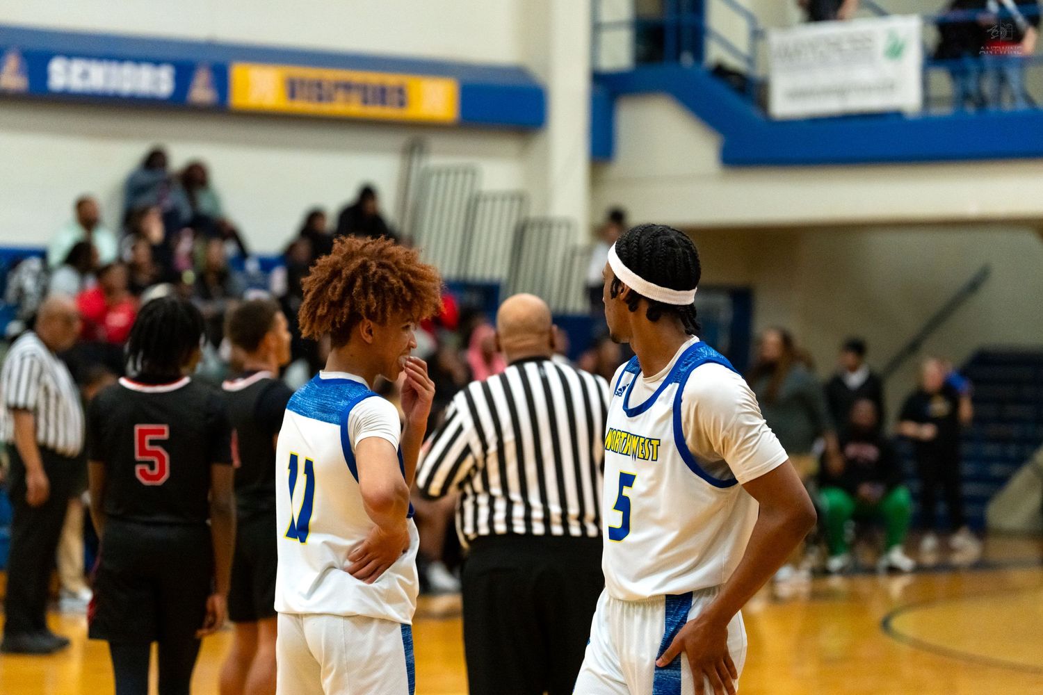 Two high school basketball players in blue and white uniforms confer near half court with referee nearby.