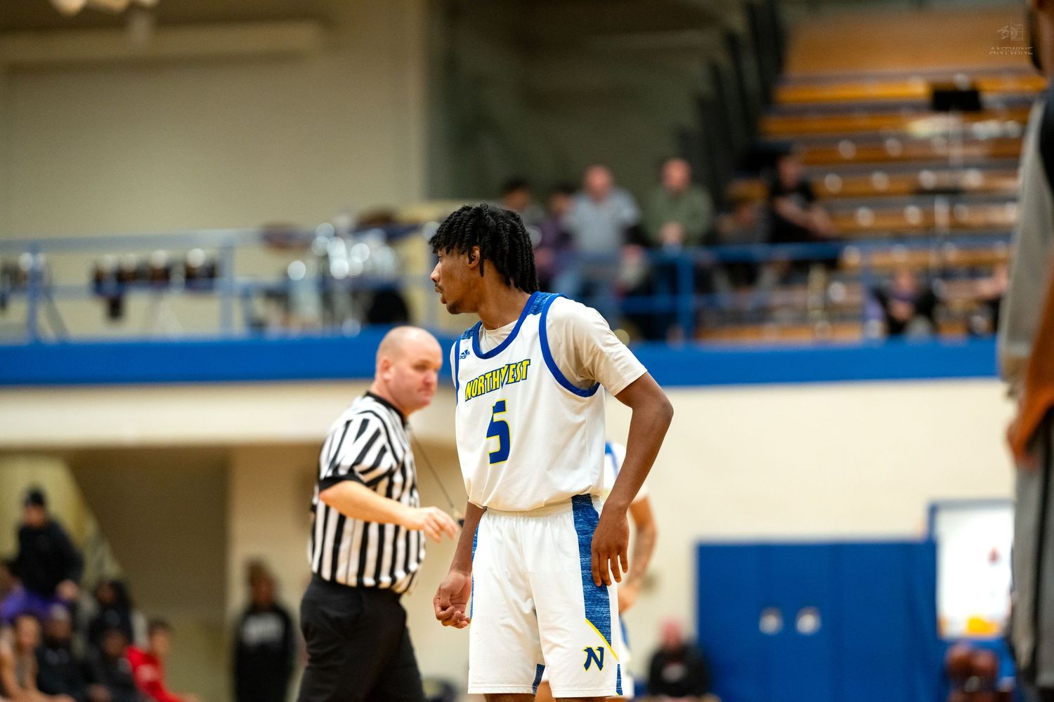 Player #5 in white and blue basketball uniform stands on court with referee in background during indoor game.