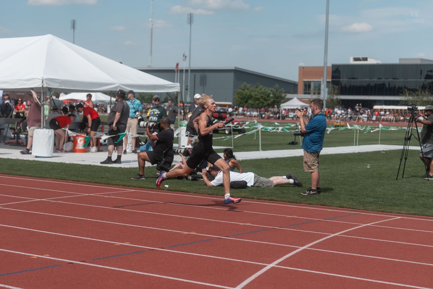 Runners sprint toward finish line at outdoor track meet as photographer captures the race on sunny day.