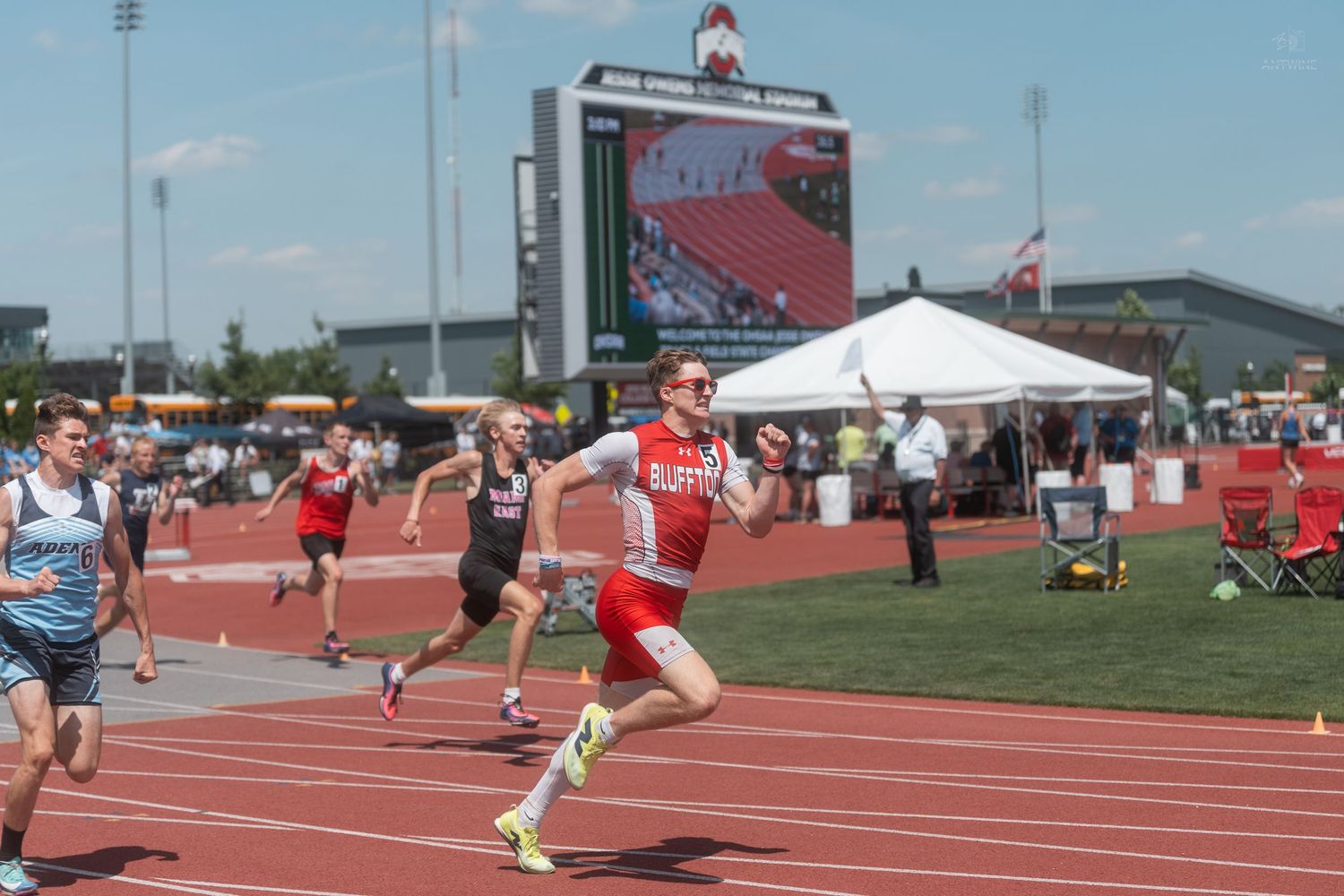 Smiling runner in red uniform sprinting near the finish line at an outdoor high school track meet.