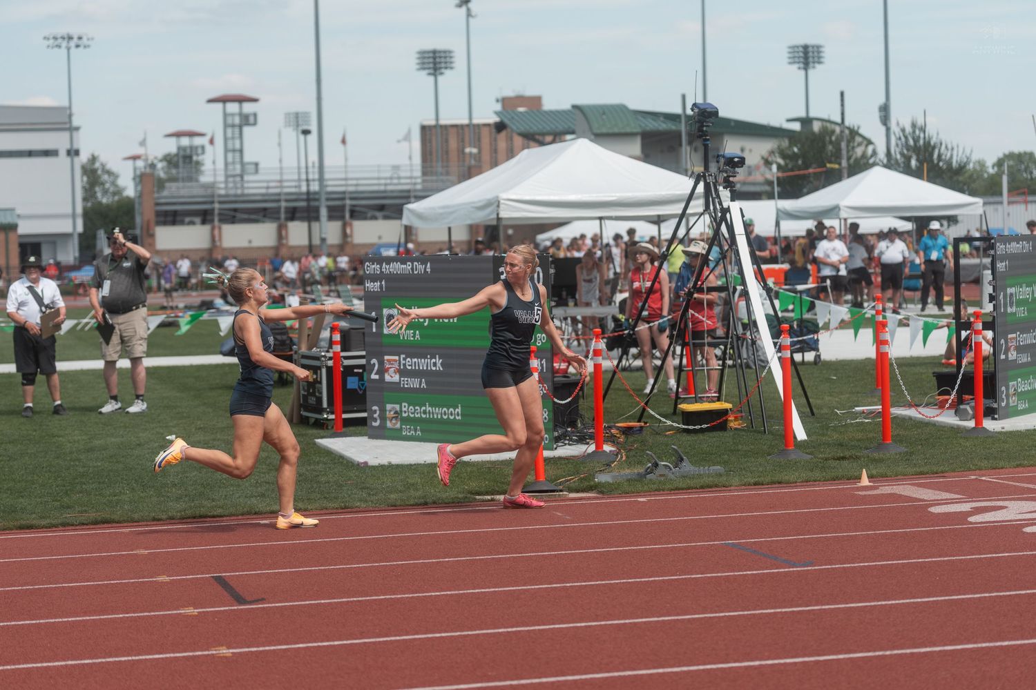 Two female athletes perform a baton exchange during a relay race at a high school state track championship.