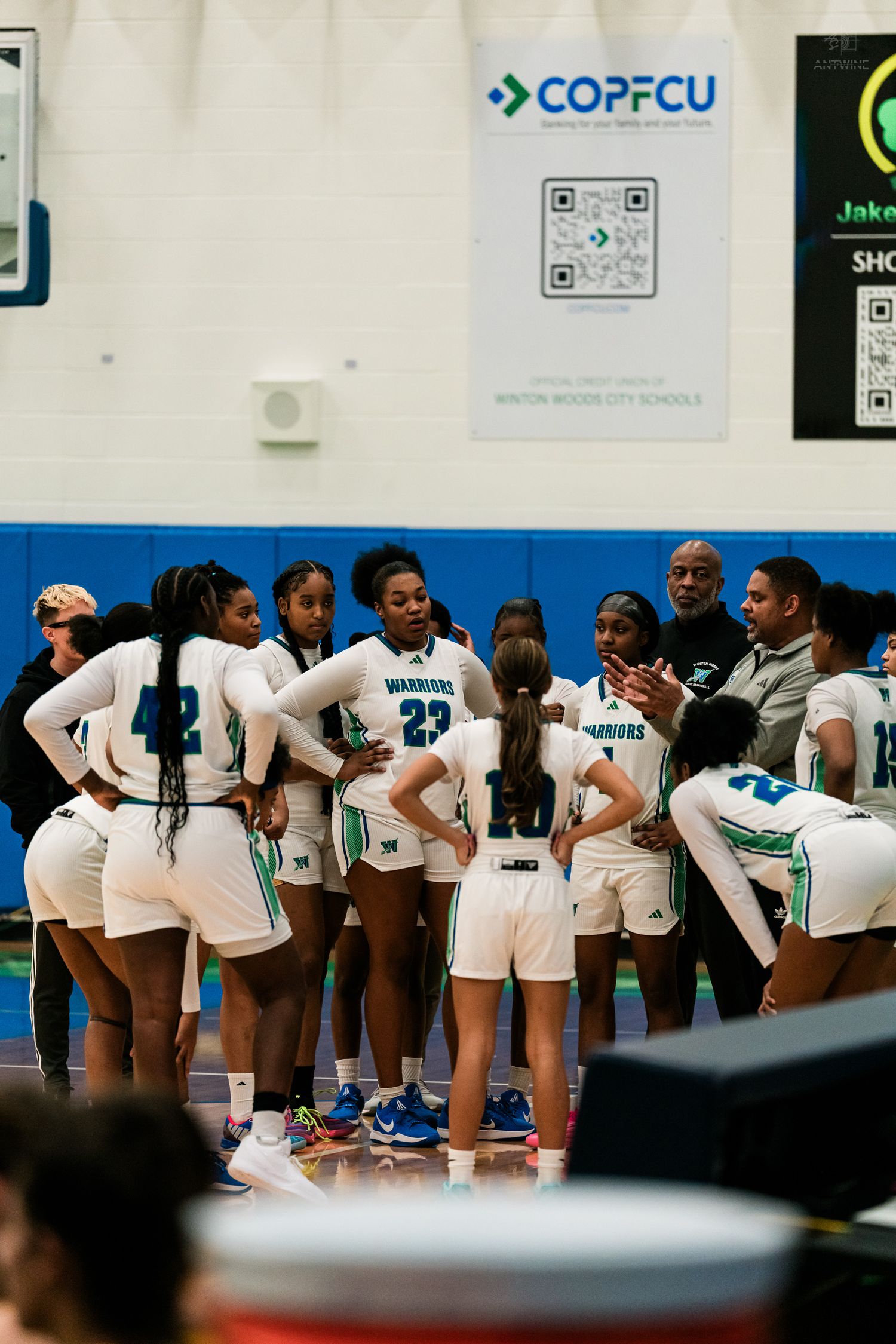 The Warriors girls basketball team gathers in a huddle with coaches during a timeout near a COPFCU banner.