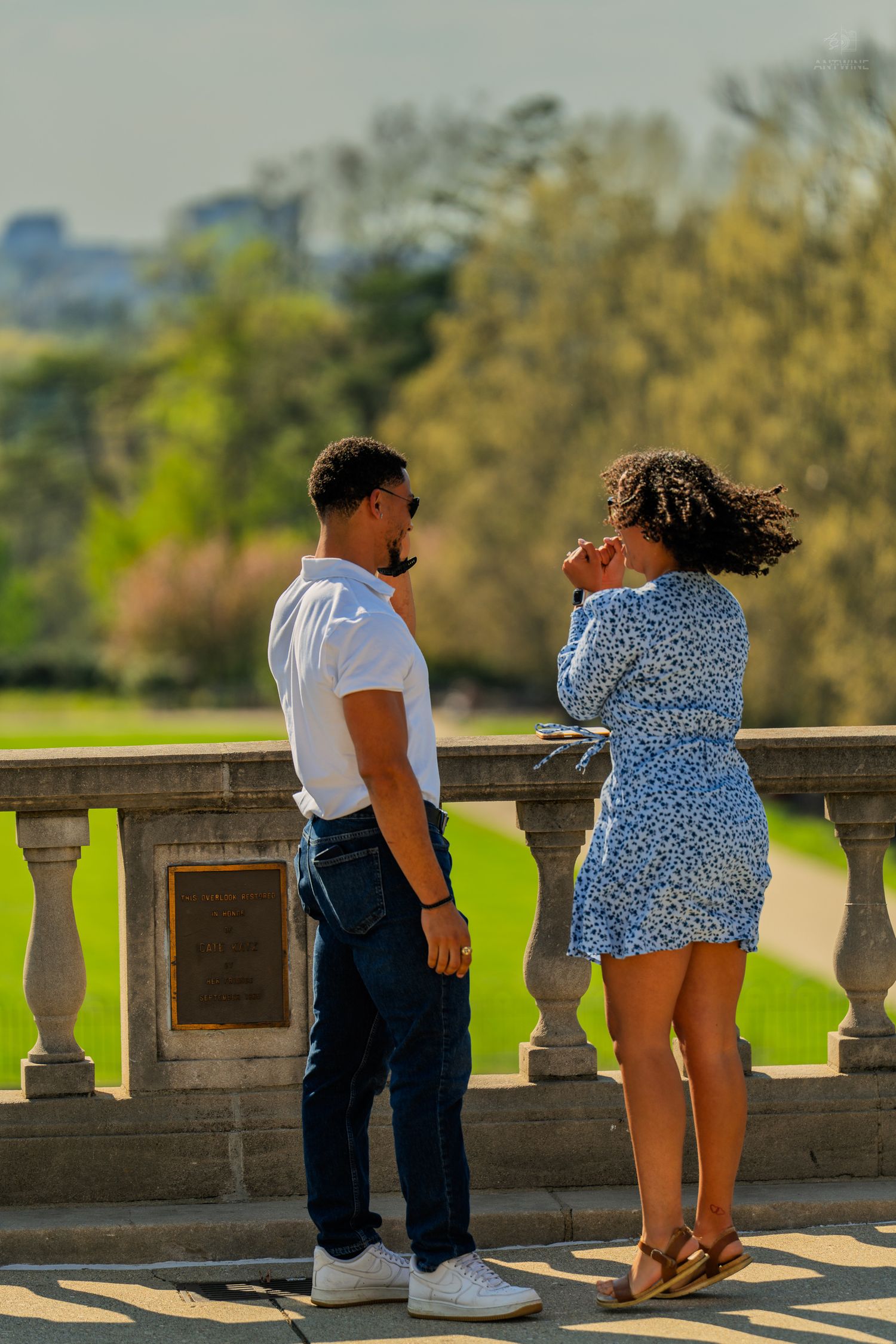 Couple standing by stone balustrade in park, woman in blue floral dress facing man in white shirt and jeans.