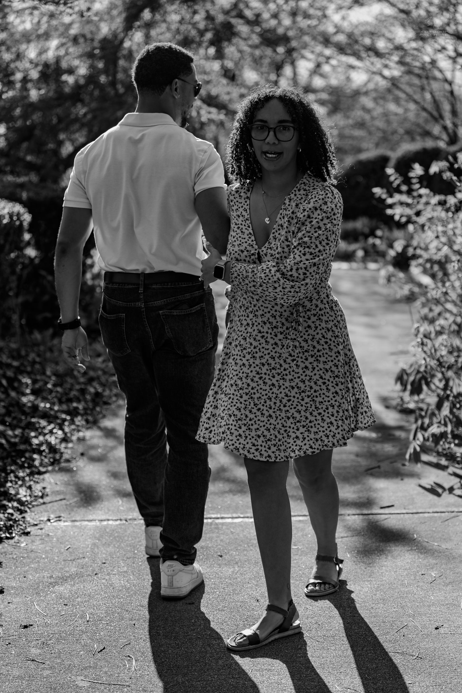 Black and white image of a curly-haired woman in a floral dress holding hands with a man in a park.