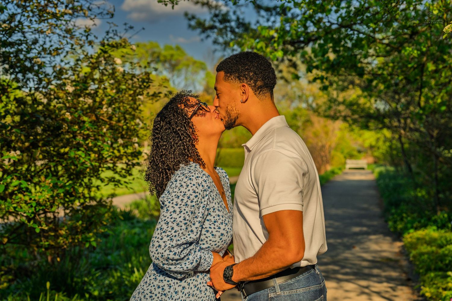 Couple sharing a tender kiss on a tree-lined park path in warm golden hour sunlight.