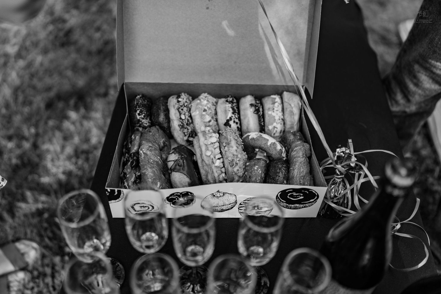 Black and white image of assorted donuts in a box with champagne flutes arranged on a dark surface outdoors.