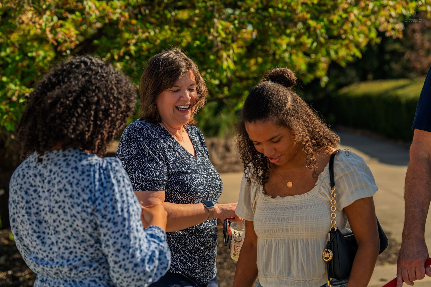Three women share a joyful moment outdoors in warm sunlight, smiling and looking at something together.