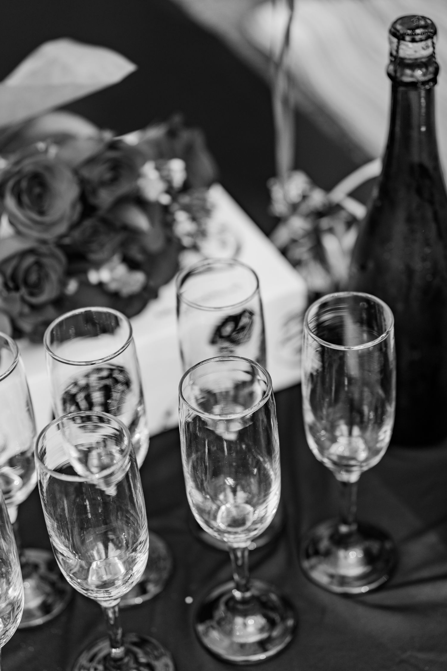 Black and white photo of champagne flutes, bottle, and rose bouquet arranged on a table.
