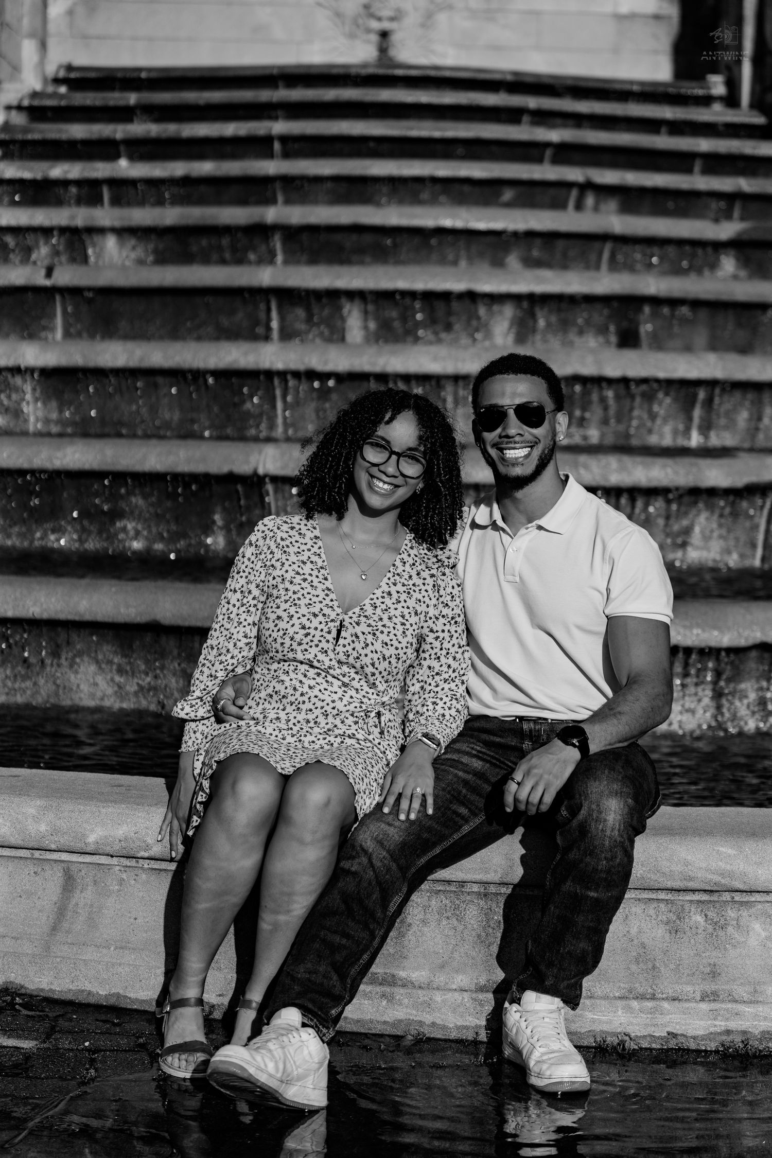 A smiling Black couple sits together on stone steps near a fountain in a black and white photo.