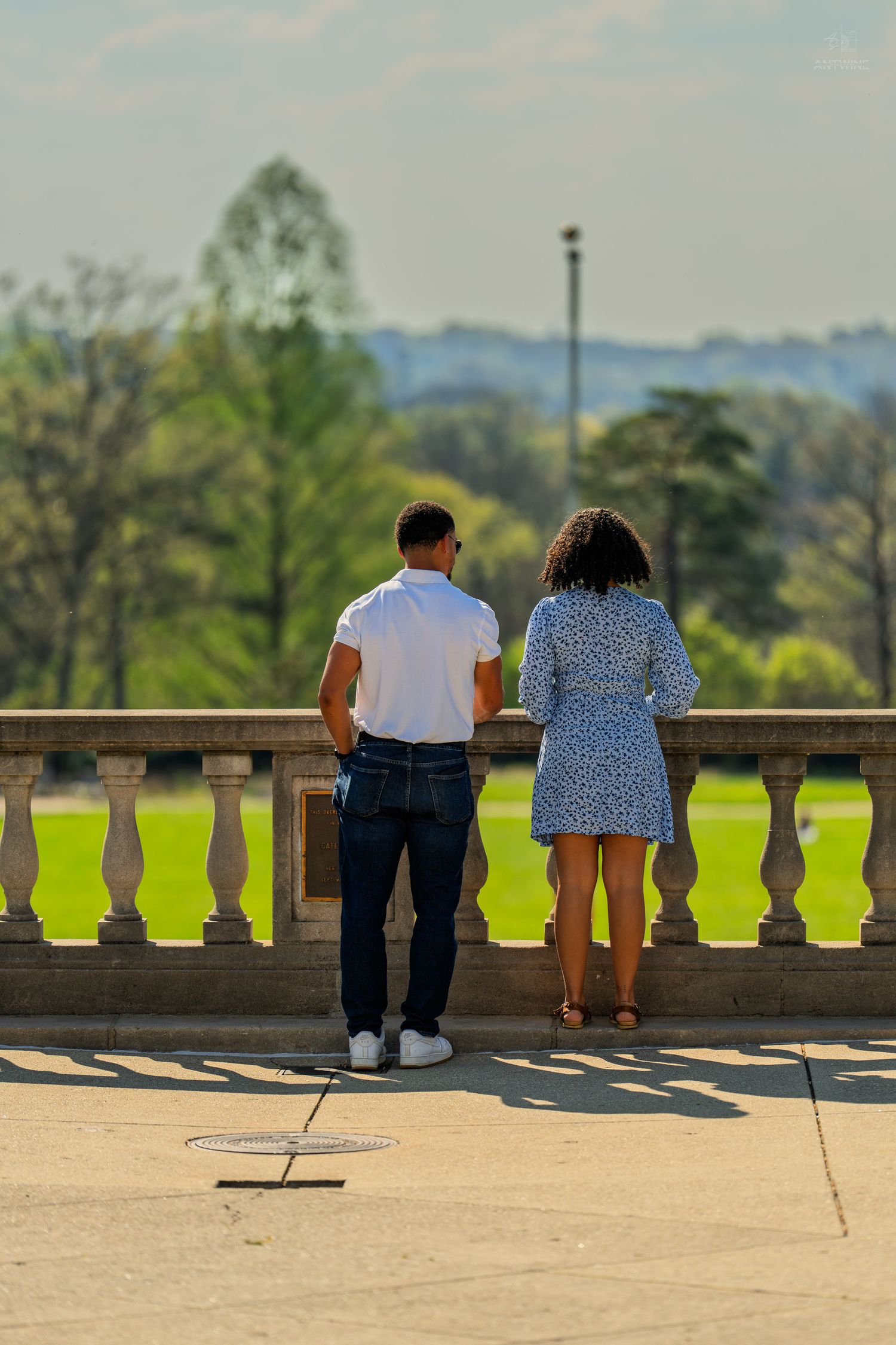 Man in white shirt and woman in floral dress admiring scenic park view from terrace.