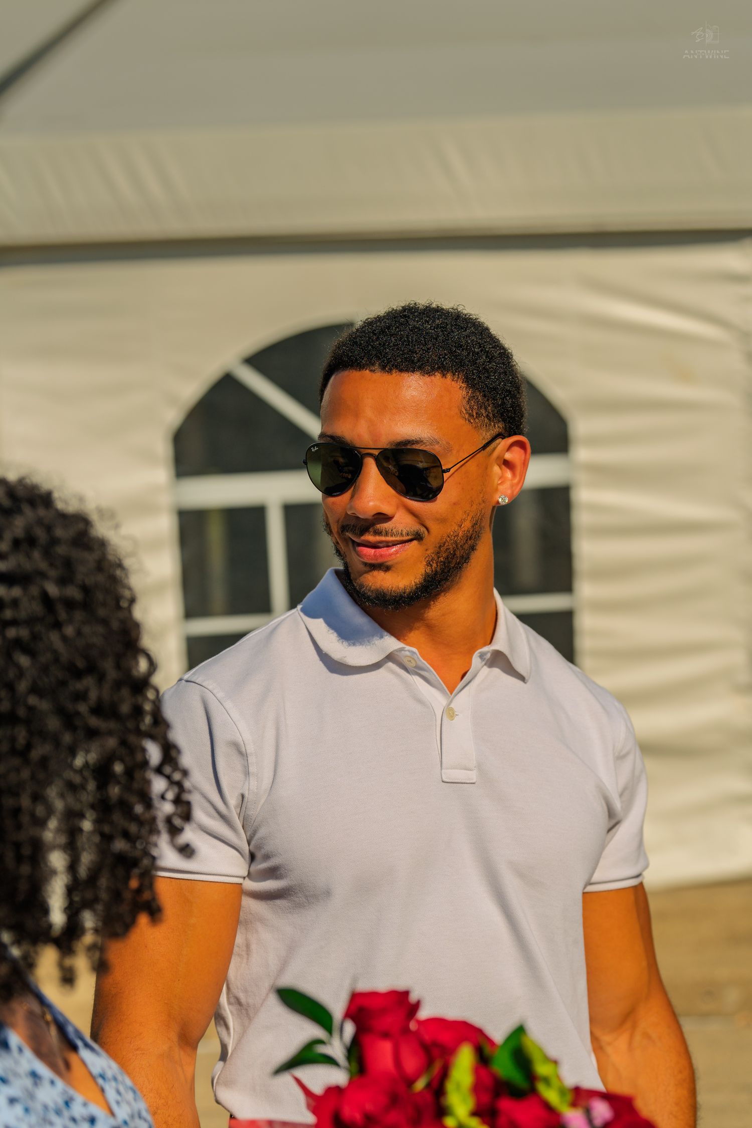 A smiling man wearing aviator sunglasses and a white polo shirt standing outdoors near a white marquee tent.