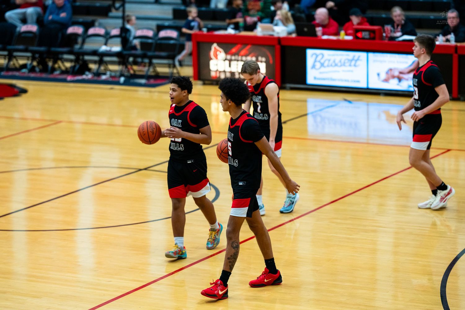 High school basketball players in black and red uniforms dribbling during a game at Bassett High School gym.