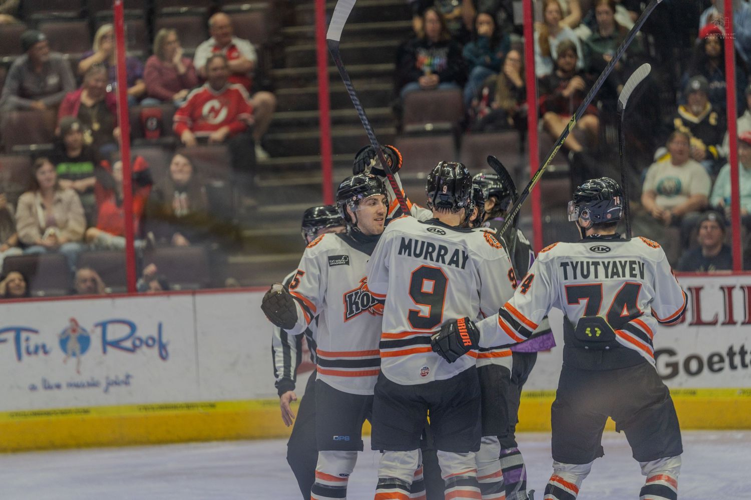 Hockey players celebrate a goal on the ice, with Murray #9 and Tyutyayev #74 visible in orange and white jerseys.