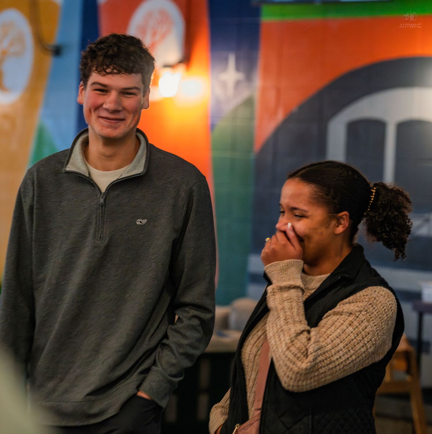 A tall young man in a gray Lacoste pullover laughs beside a smiling woman in a black vest at an indoor event.