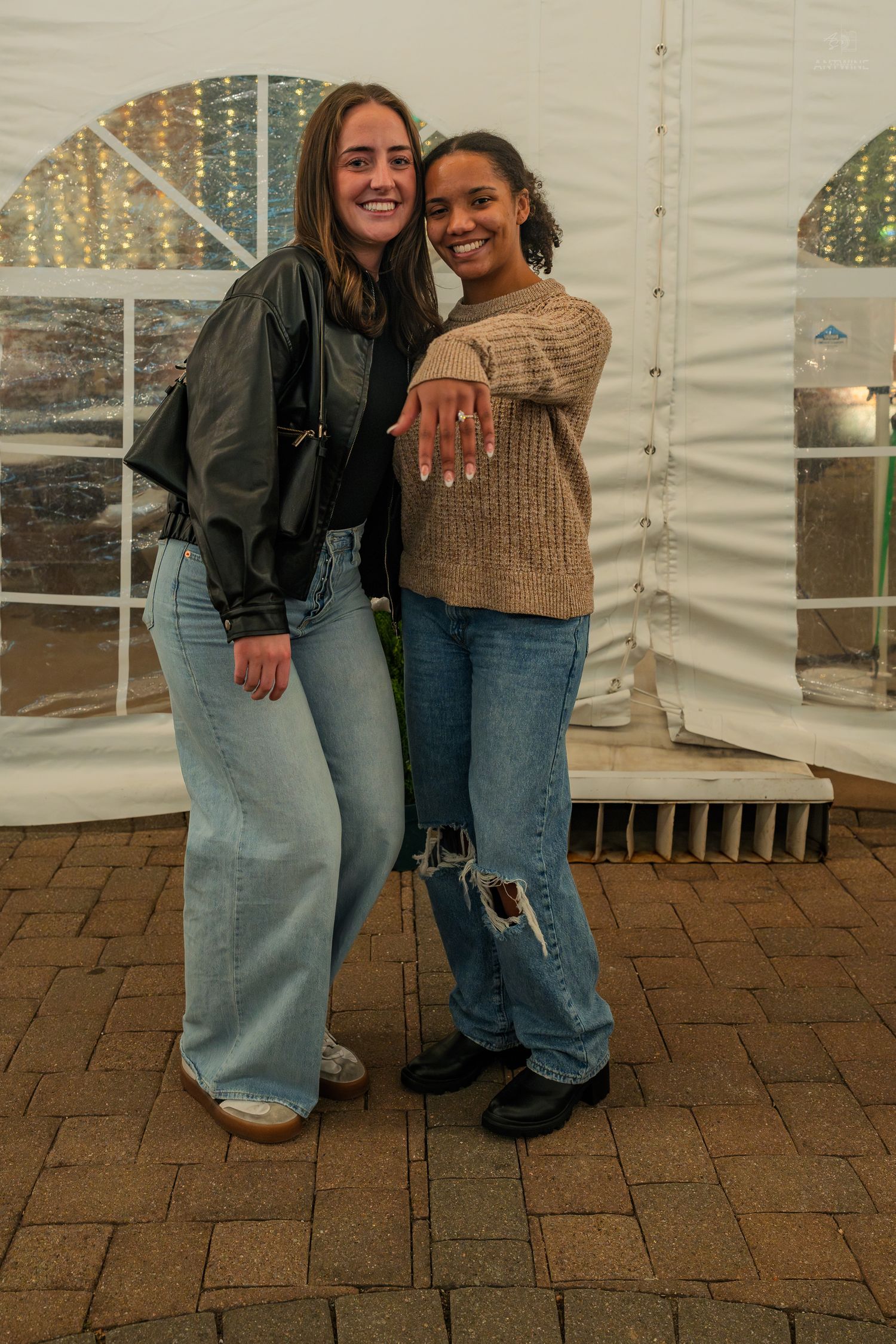 Two young women smiling and posing together inside a white tent, one displaying an engagement ring.