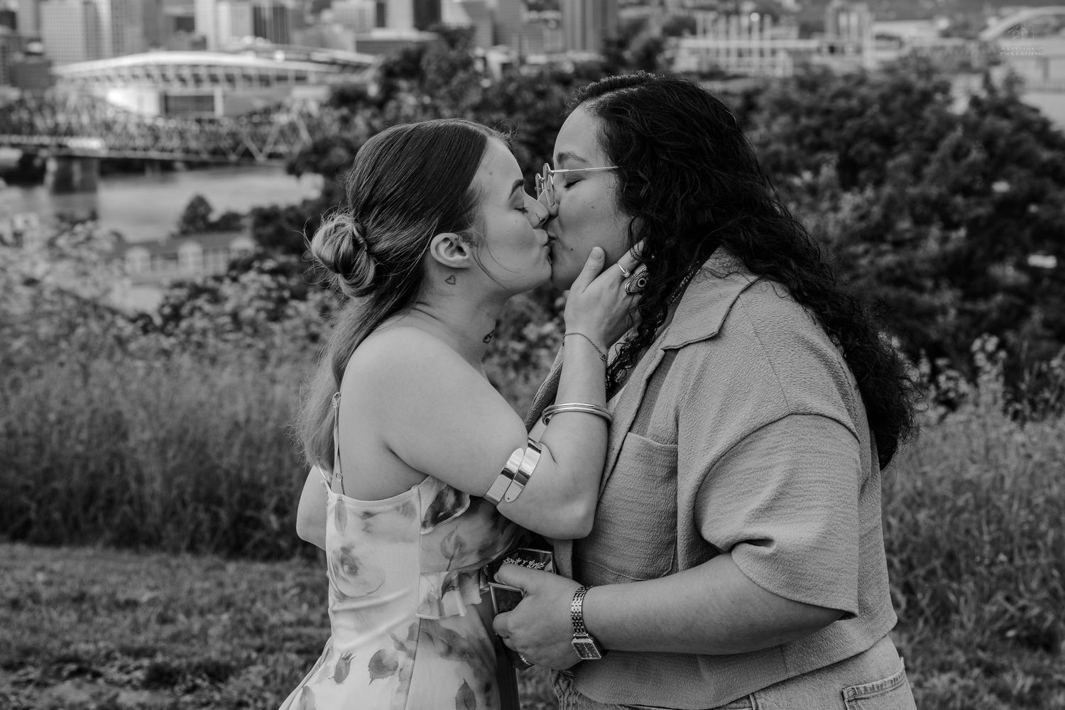 Black and white photo of two women kissing in a park with a city skyline visible in the background.