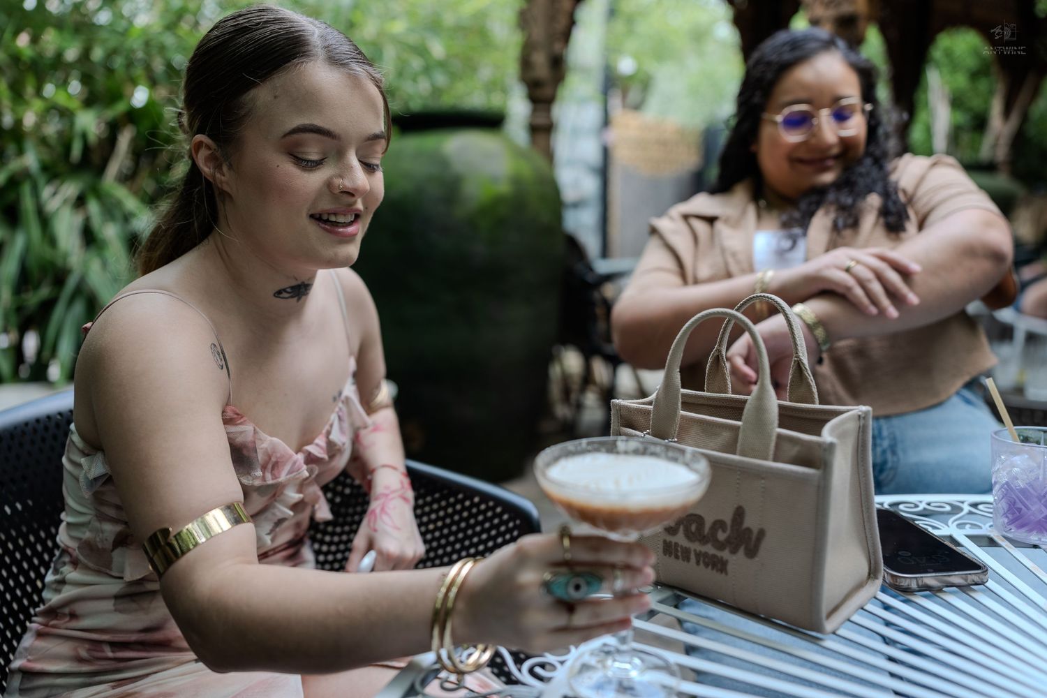 Two women smiling at outdoor cafe table with cocktail and designer tote bag.