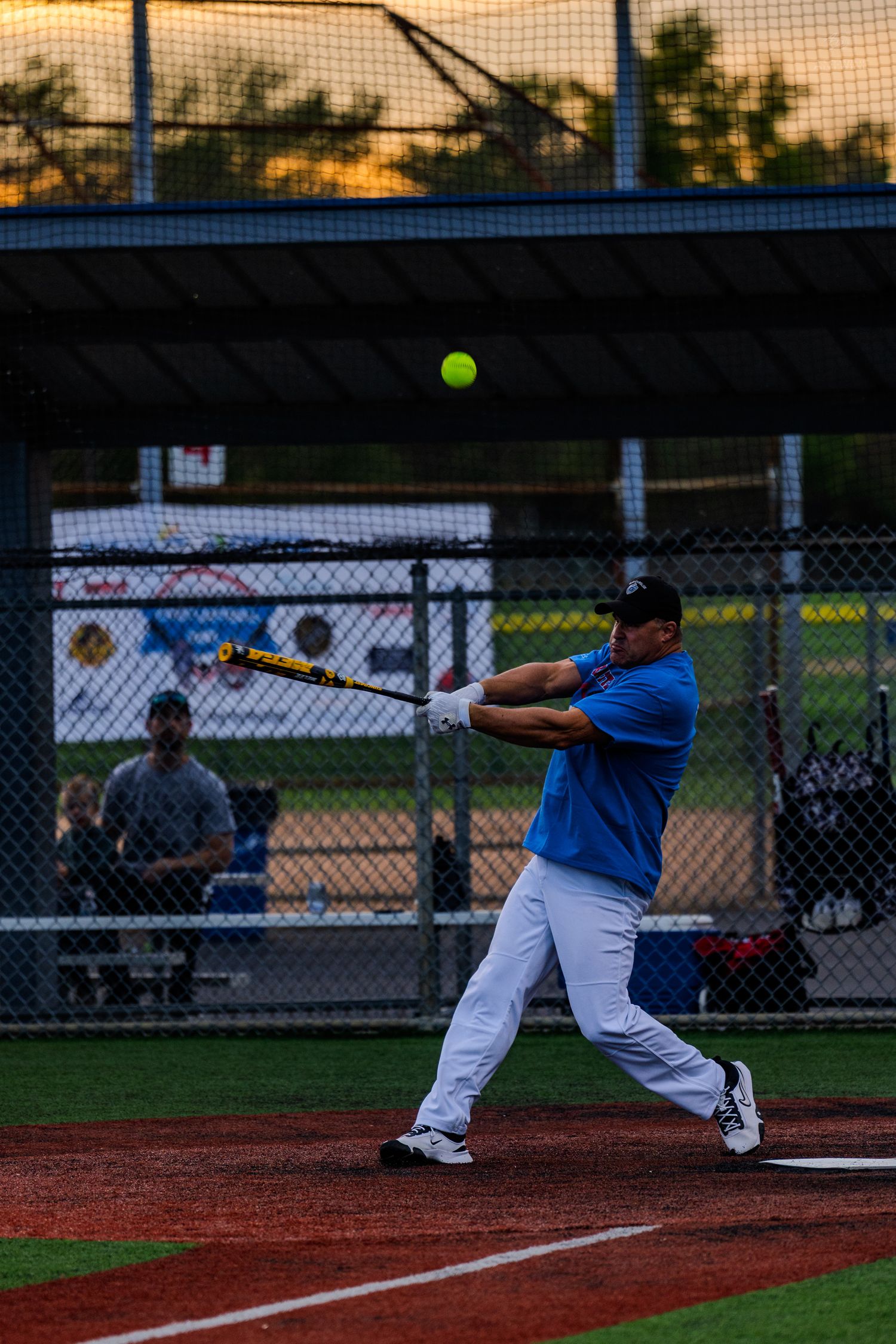 Softball batter in blue jersey making contact with a yellow ball during an evening recreational league game.