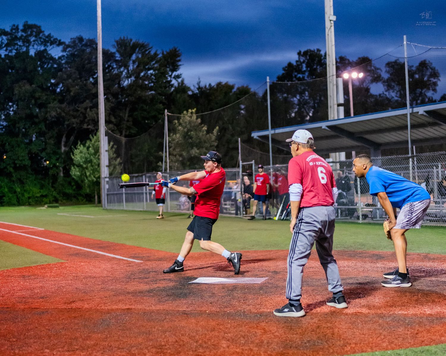 Night softball game with batter in red, catcher, and umpire on a lit field under a dramatic dusk sky.