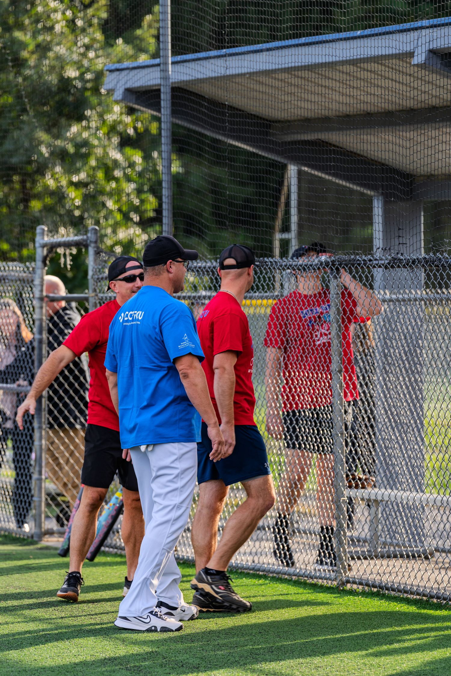 Men in red and blue shirts walk near a chain-link fence at an outdoor baseball field during a sunny day.